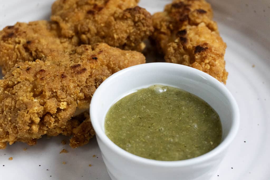 Four pieces of crispy fried chicken are arranged on a white plate next to a small white bowl filled with green dipping sauce.