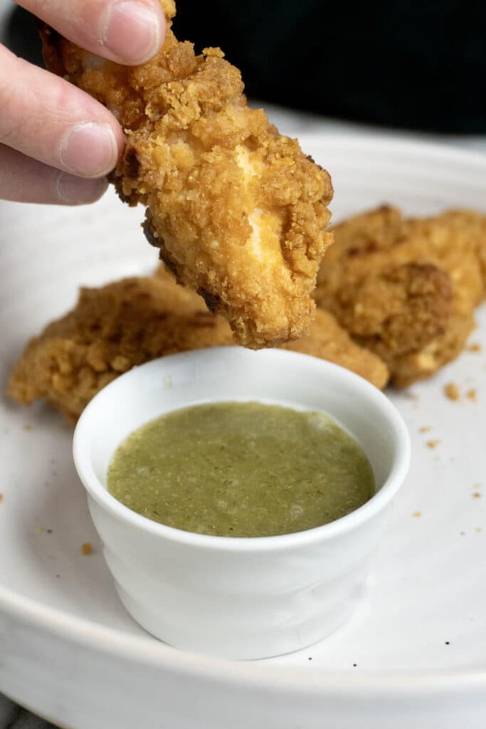 A hand holds a piece of crispy fried chicken above a small white bowl filled with green dipping sauce, with more fried chicken pieces on a white plate in the background.