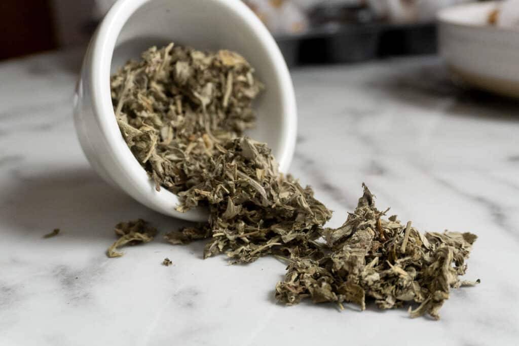 Dried sage leaves spill out of a small white ceramic cup onto a marble surface.