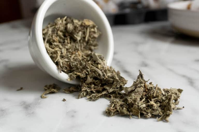 Dried sage leaves spill out of a small white ceramic cup onto a marble surface.