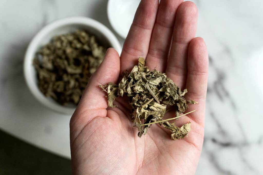 A hand holding a small pile of dried, crumbled herbs, with a white bowl of more dried herbs in the blurred background on a marble surface.