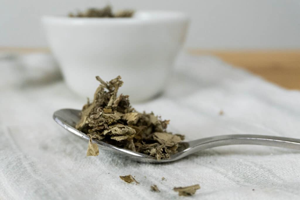 A close-up of dried tea leaves on a metal spoon, with a white bowl filled with more tea leaves blurred in the background, all resting on a light-colored cloth.