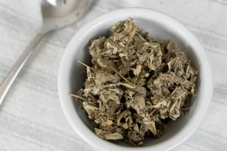 A white bowl filled with dried, crumbled herbs is placed on a light-colored fabric surface, with a metal spoon lying next to it.
