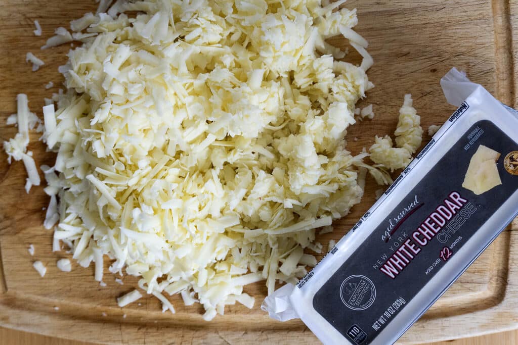 A pile of freshly grated white cheddar cheese sits on a wooden cutting board next to an unopened block of white cheddar cheese in its packaging.