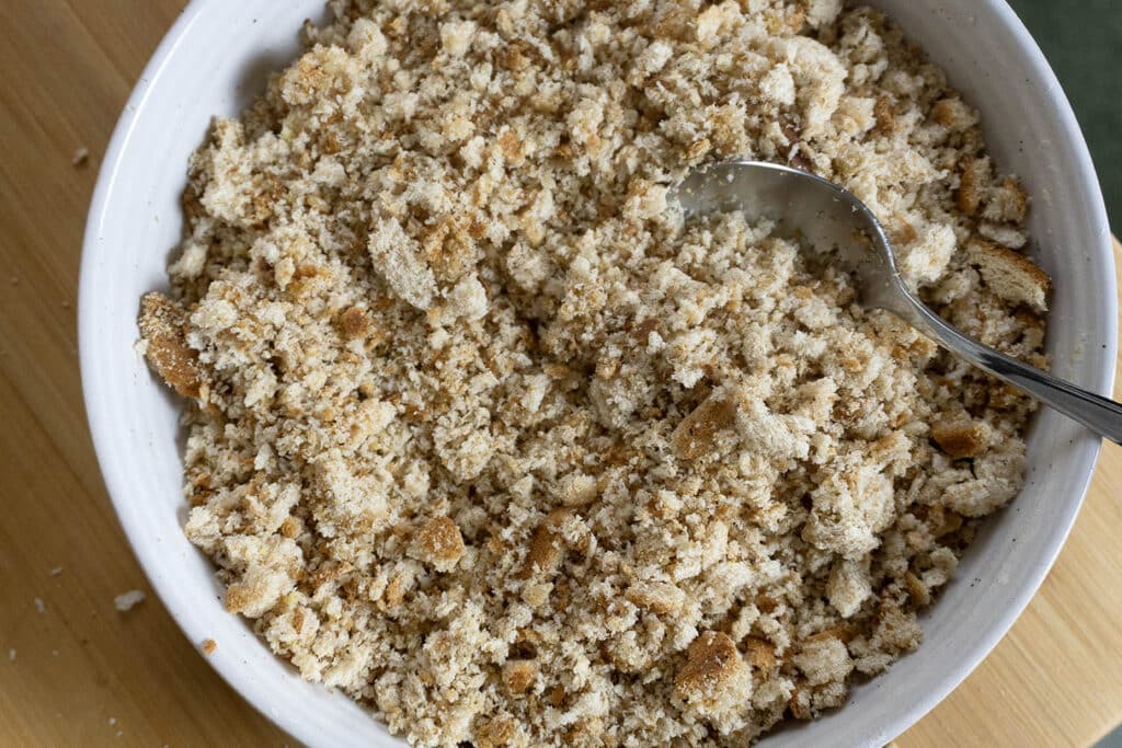 A white bowl filled with coarse, crumbled bread crumbs sits on a wooden surface. A metal spoon rests inside the bowl.
