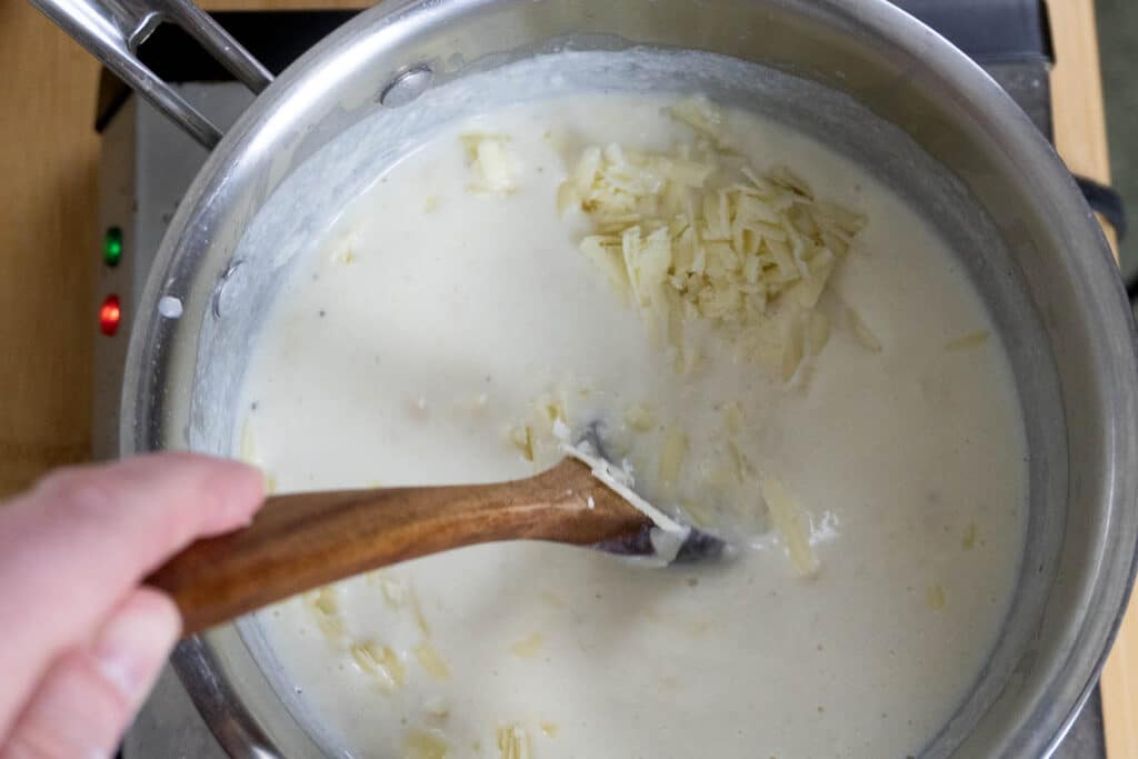 A hand stirs a creamy white sauce with shredded cheese in a saucepan on a stove using a wooden spoon.