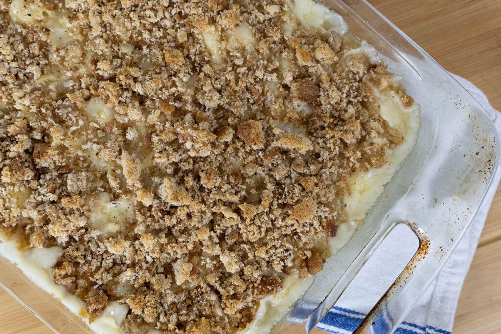 A glass baking dish filled with a creamy casserole topped with a golden brown breadcrumb and crouton crust, sitting on a wooden surface next to a white and blue striped towel.