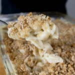A close-up of a spoonful of creamy baked macaroni and cheese topped with golden, crispy breadcrumbs, held above a glass baking dish filled with the same macaroni and cheese.