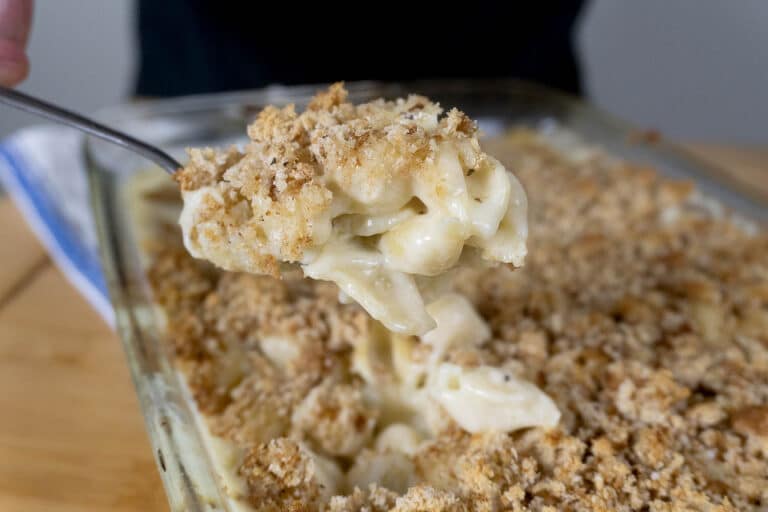 A close-up of a spoonful of creamy baked macaroni and cheese topped with golden, crispy breadcrumbs, held above a glass baking dish filled with the same macaroni and cheese.