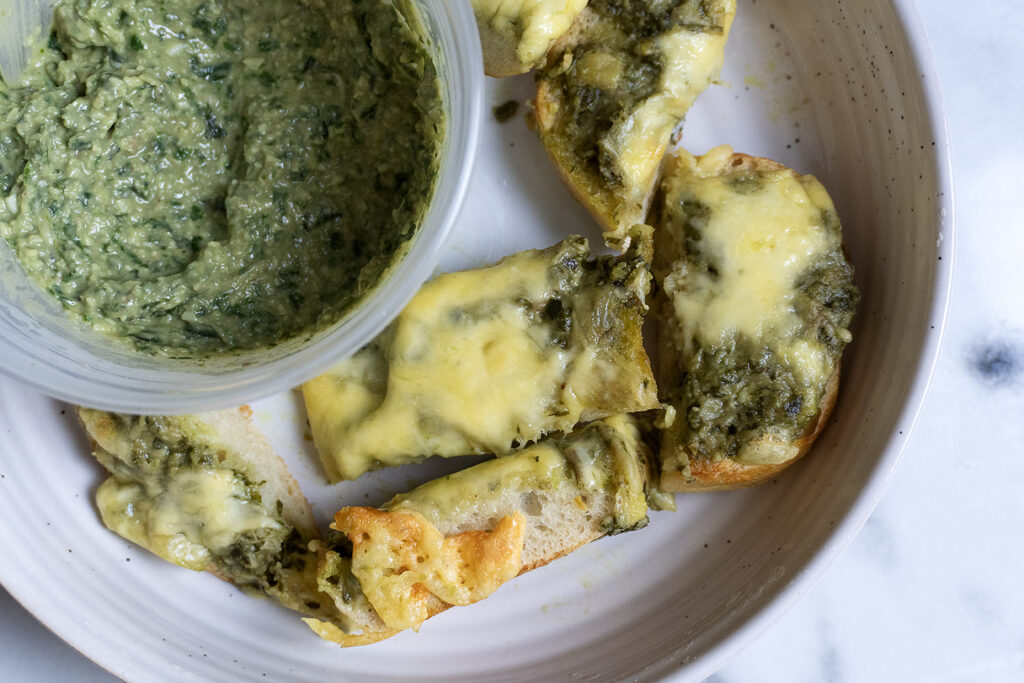 A plate with slices of bread topped with melted cheese and green pesto, next to a container of green dip or spread. The plate is on a white marble surface.