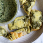 A plate with slices of bread topped with melted cheese and green pesto, next to a container of green dip or spread. The plate is on a white marble surface.