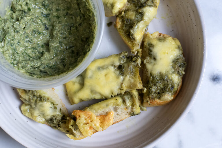 A plate with slices of bread topped with melted cheese and green pesto, next to a container of green dip or spread. The plate is on a white marble surface.