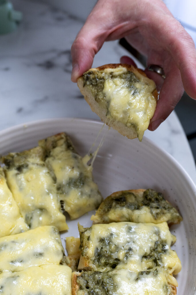 A hand lifts a slice of cheesy bread topped with melted cheese and green pesto from a plate with several similar slices. The cheese stretches as the slice is pulled away.