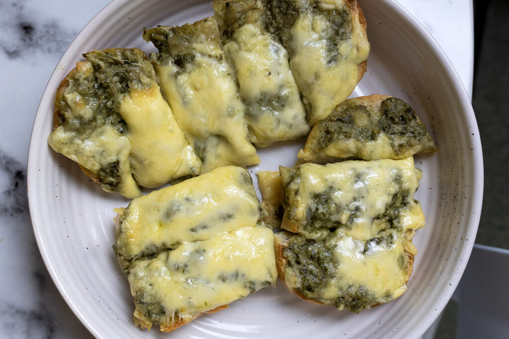 Slices of bread topped with green pesto and melted cheese are arranged on a white plate, set on a marble surface.