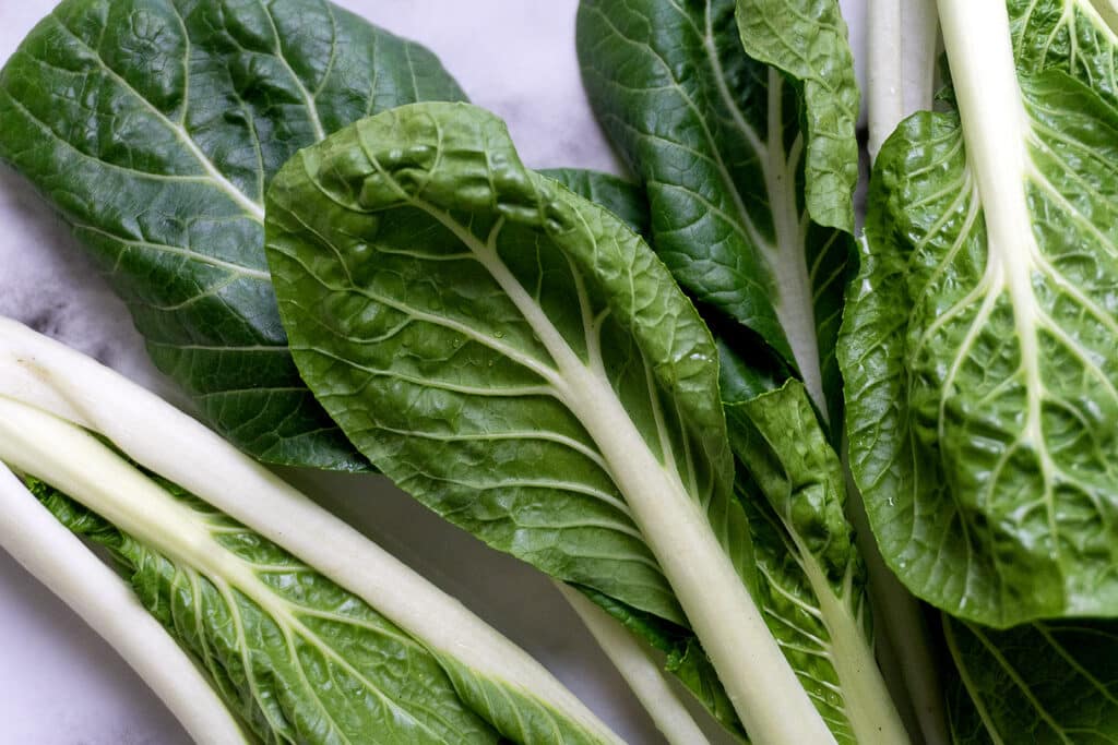 Several fresh green bok choy leaves with crisp white stems are spread out on a light-colored surface. The leaves have visible veins and a vibrant, healthy appearance.