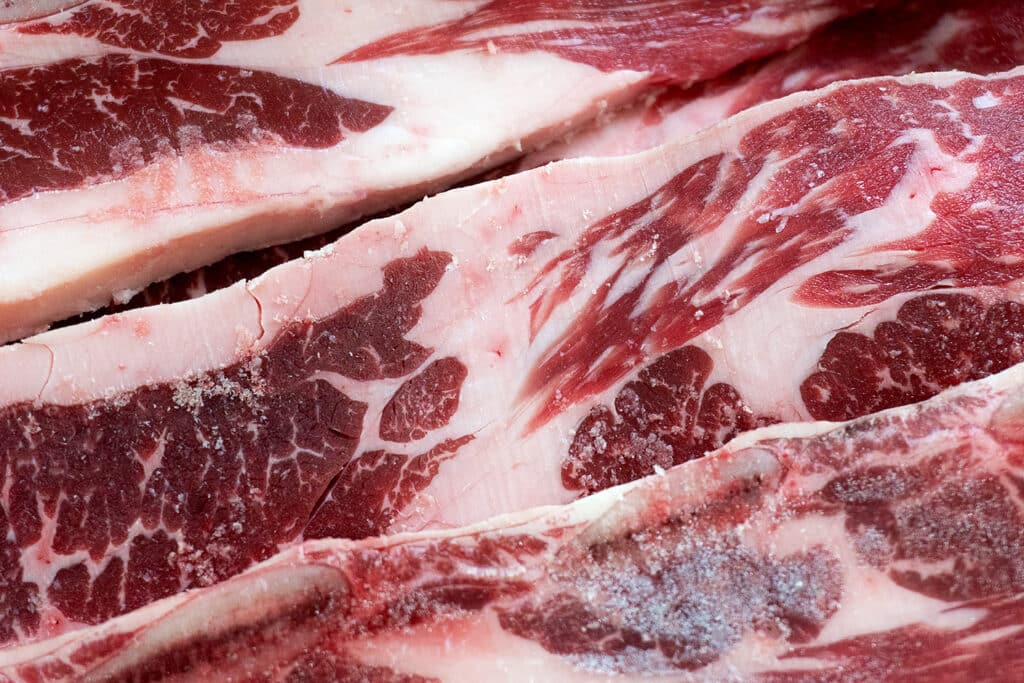 Close-up of raw, marbled beef slices with visible fat and muscle fibers. The meat appears fresh with some frost on the surface, suggesting it may be partially frozen.