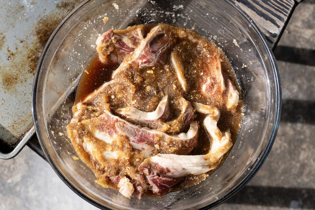Raw ribs marinating in a glass bowl with a brown, grainy sauce, likely containing spices and seasonings, sitting on a kitchen counter in natural light.