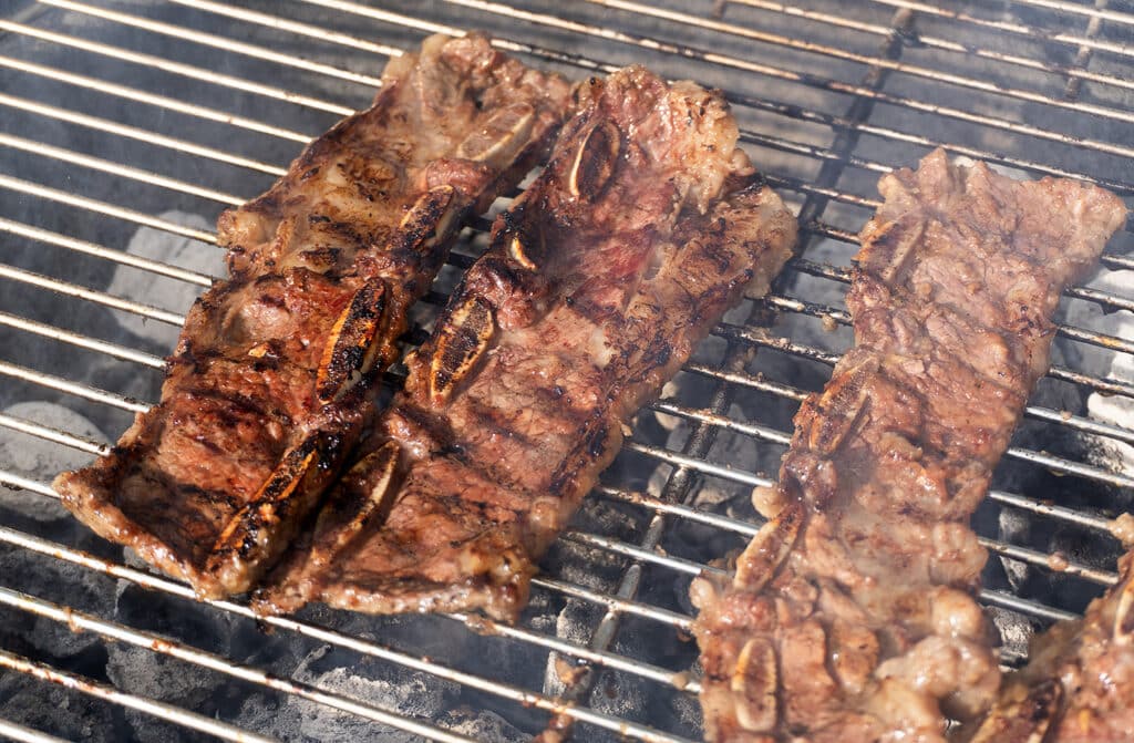 Three pieces of marinated beef short ribs are grilling over hot charcoal on a metal barbecue grill, with visible grill marks and sizzling juices.