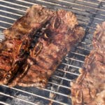 Three pieces of marinated beef short ribs are grilling over hot charcoal on a metal barbecue grill, with visible grill marks and sizzling juices.