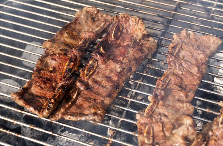 Three pieces of marinated beef short ribs are grilling over hot charcoal on a metal barbecue grill, with visible grill marks and sizzling juices.
