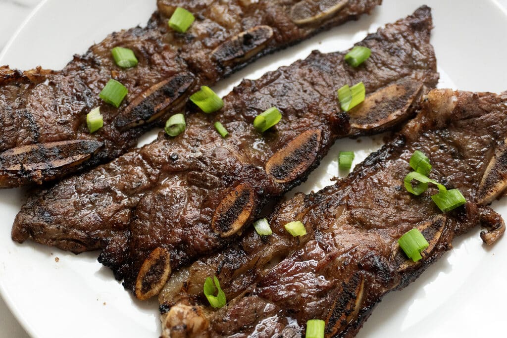 Grilled short ribs garnished with chopped green onions are arranged on a white plate. The ribs are browned with visible grill marks and bone sections.