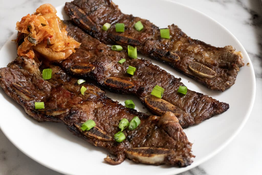 Grilled Korean short ribs garnished with chopped green onions, served on a white plate with a side of kimchi.