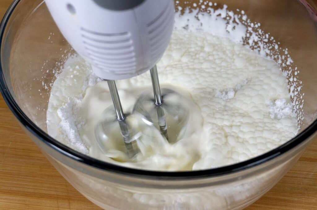 A close-up of an electric hand mixer whipping cream in a glass bowl on a wooden surface. The beaters are blending the cream, creating a frothy texture.