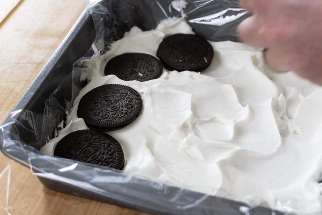 A hand places chocolate sandwich cookies on a layer of whipped cream in a plastic-lined square baking pan on a wooden surface.