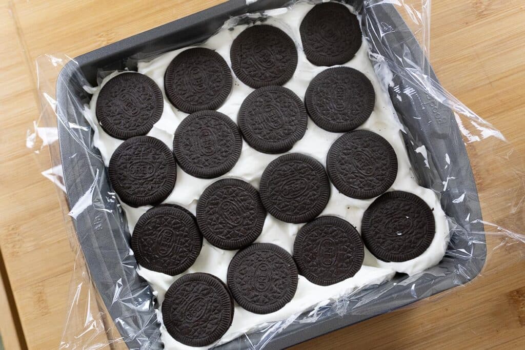 A baking pan lined with plastic wrap contains a layer of whole Oreo cookies arranged on top of white cream filling, ready to be chilled or set on a wooden surface.