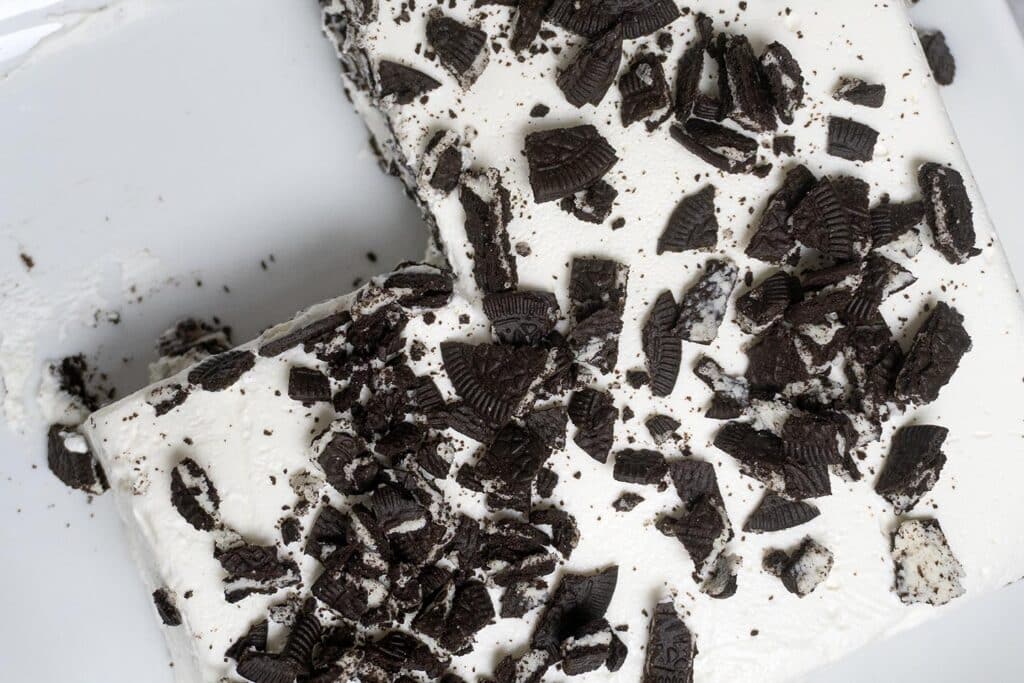 A rectangular cake with white frosting topped with crumbled chocolate sandwich cookies, with a corner slice removed, on a white surface.