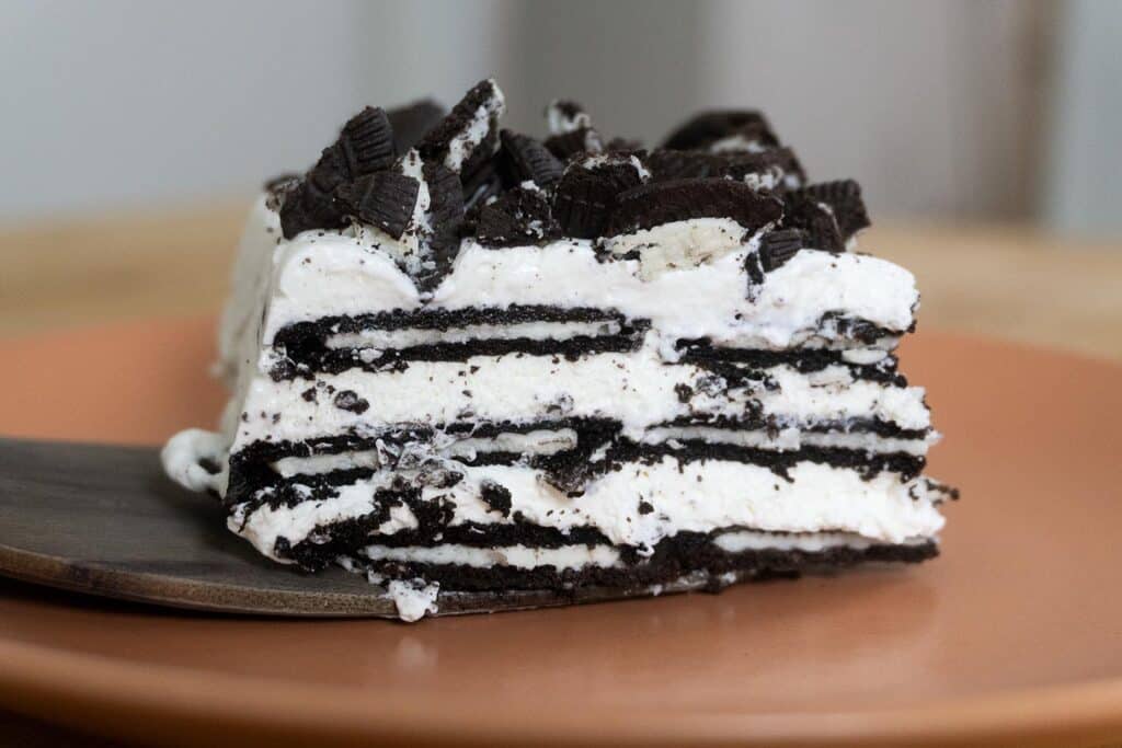 A close-up of a slice of cookies and cream icebox cake with layers of chocolate cookies and whipped cream, topped with crumbled cookies, served on a brown plate.