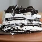 A close-up of a slice of cookies and cream icebox cake with layers of chocolate cookies and whipped cream, topped with crumbled cookies, served on a brown plate.