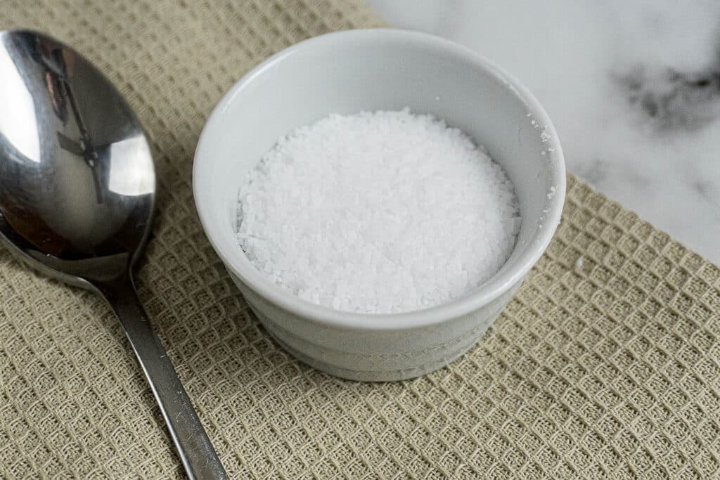 A small white ramekin filled with flaky sea salt sits on a beige textured cloth next to a metal spoon.