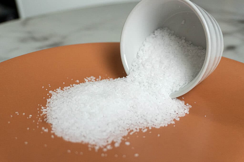 A small white bowl tipped over on an orange surface, spilling coarse white salt crystals onto the plate.