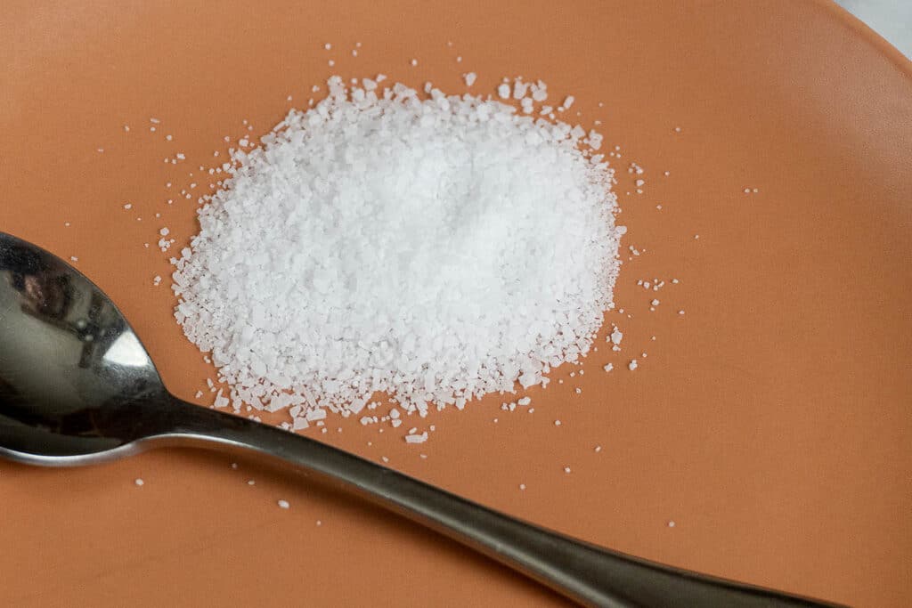 A small pile of coarse white salt is on an orange plate, with a metal spoon placed beside it.