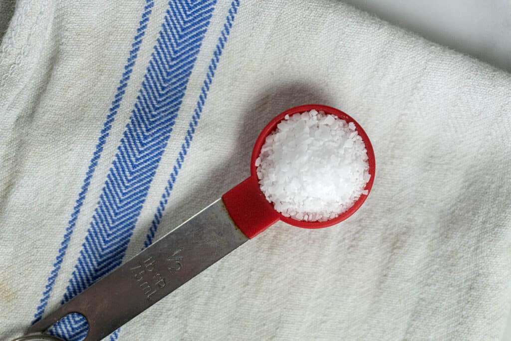 A red 1/2 tablespoon measuring spoon filled with coarse salt rests on a white cloth with blue stripes.