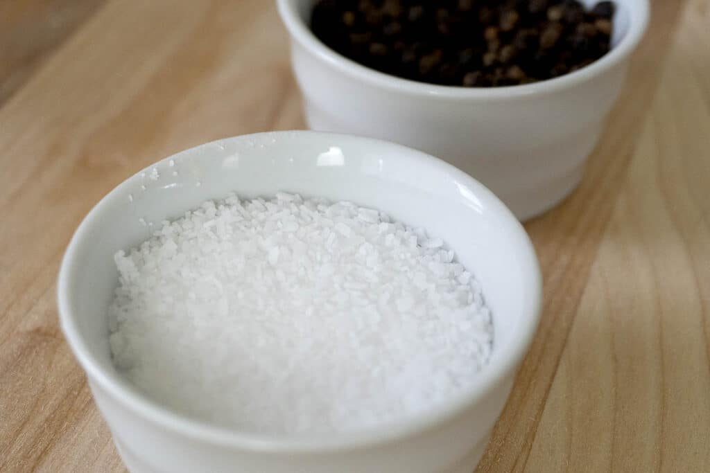 A small white bowl filled with coarse salt sits in the foreground, with another white bowl containing whole black peppercorns slightly out of focus in the background on a wooden surface.