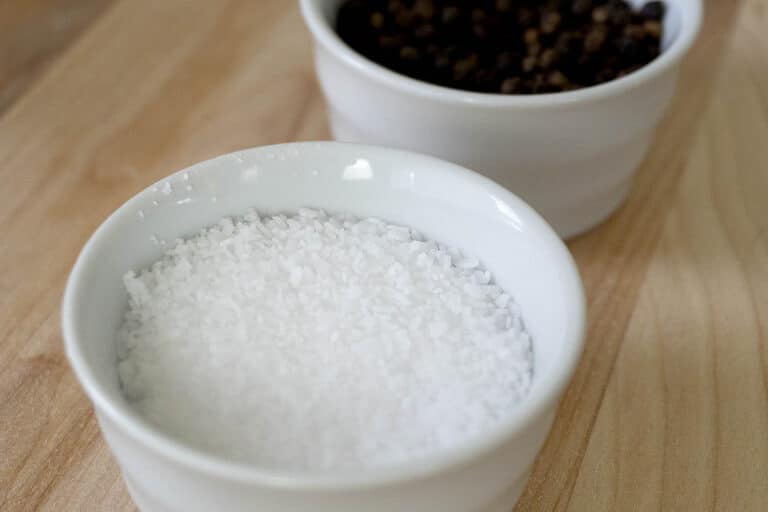 A small white bowl filled with coarse salt sits in the foreground, with another white bowl containing whole black peppercorns slightly out of focus in the background on a wooden surface.