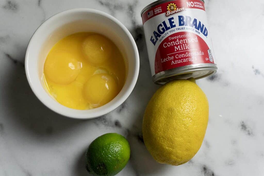 A bowl with cracked eggs, a can of Eagle Brand sweetened condensed milk, a lemon, and a lime are arranged on a marble surface.