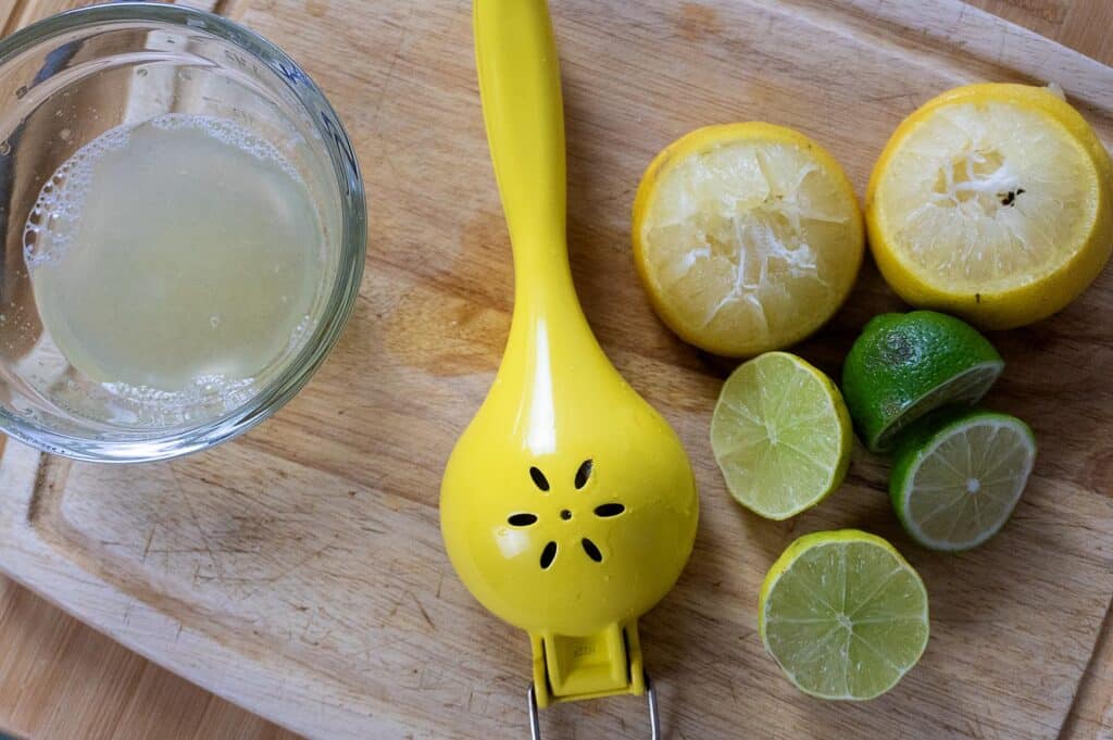 A glass of juice, a yellow citrus squeezer, and several halved lemons and limes are arranged on a wooden cutting board. Some citrus halves appear juiced, while others are fresh and unjuiced.