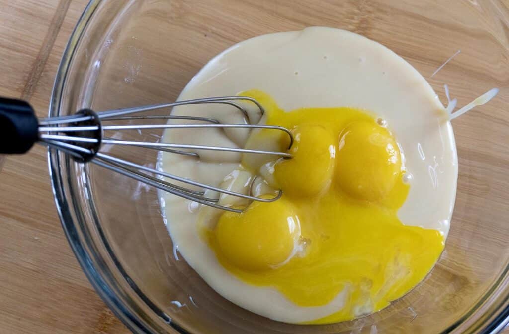 A glass bowl contains three raw eggs and a creamy liquid, with a metal whisk resting inside, ready for mixing on a wooden surface.