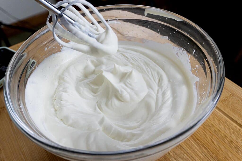 A glass bowl filled with freshly whipped cream showing soft peaks, with a hand mixer resting above the bowl. The bowl is placed on a wooden surface.