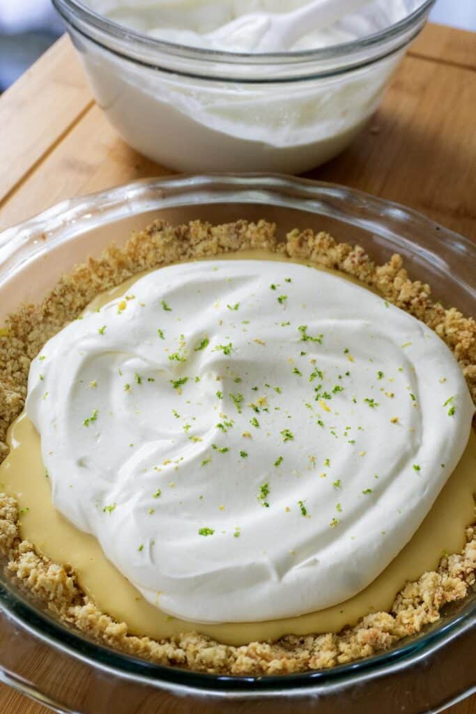 A glass pie dish filled with a key lime pie topped with whipped cream and lime zest, resting on a wooden surface. In the background, a glass bowl with more whipped cream is visible.