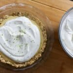 A key lime pie with a crumbly crust topped with whipped cream and lime zest sits on a wooden surface next to a bowl of whipped cream with a spatula.