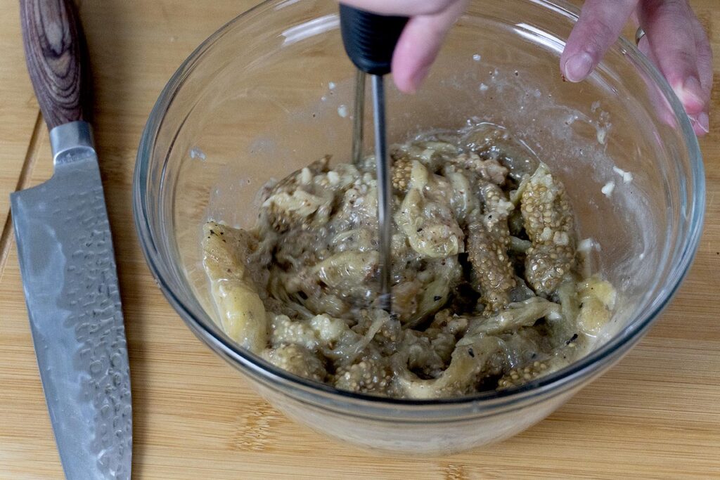 A person mashes roasted eggplant in a glass bowl with a fork or whisk on a wooden surface; a knife rests nearby.