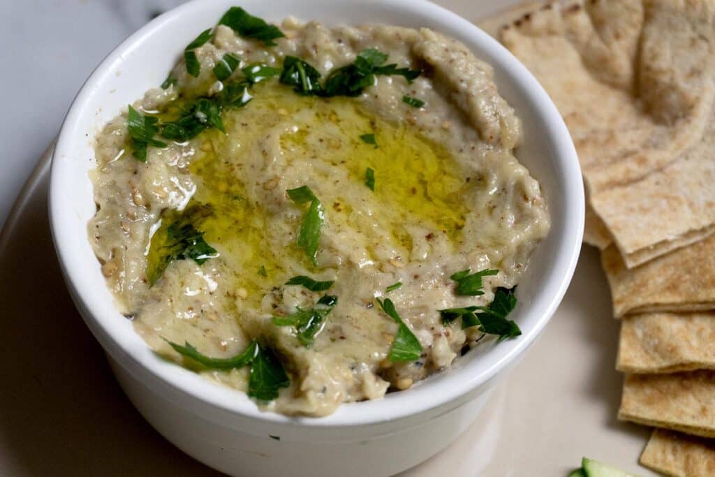 A bowl of baba ganoush topped with olive oil and chopped parsley, served with slices of pita bread on the side.