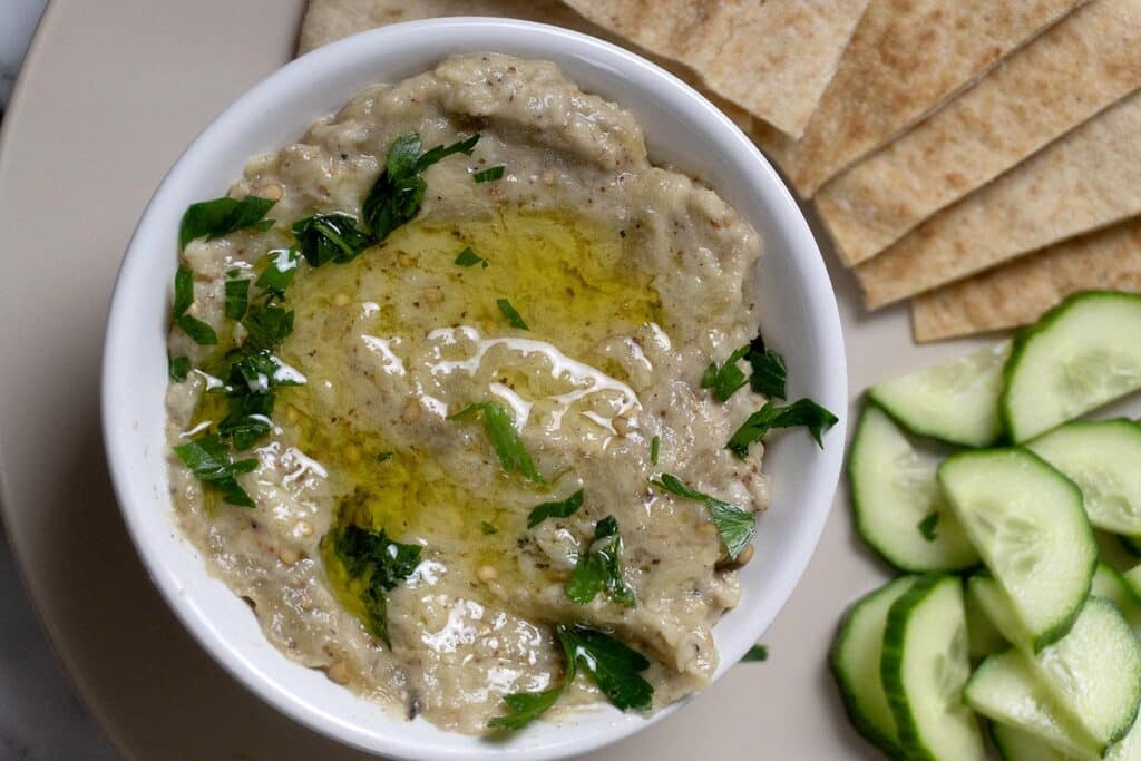 A bowl of baba ganoush topped with olive oil and chopped parsley, served with slices of pita bread and cucumber on a plate.
