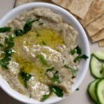 A bowl of baba ganoush topped with olive oil and chopped parsley, served with slices of pita bread and cucumber on a plate.