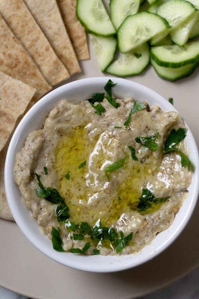 A bowl of creamy baba ganoush topped with olive oil and chopped parsley, served with sliced pita bread and cucumber rounds on a plate.