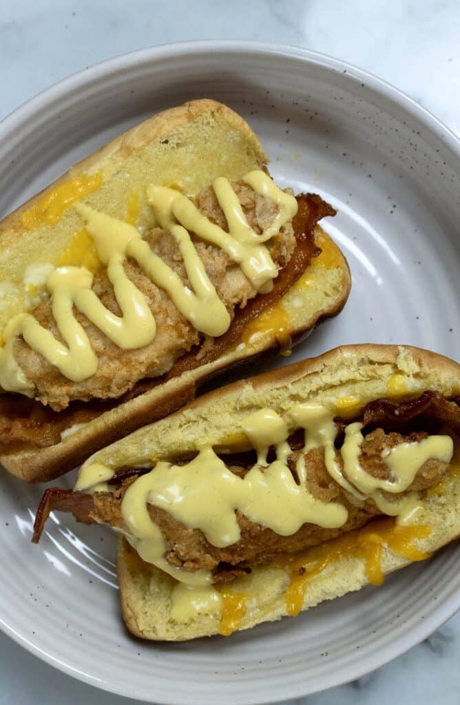 Two sandwiches on a white plate, each with a breaded fried chicken fillet, crispy bacon, melted cheese, and a generous drizzle of creamy yellow sauce in a toasted bun.